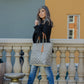 Woman wearing a black blazer, blue jeans, and heels holding a woven brown and white chevron woven bag near a railing in front of teal and yellow painted buildings