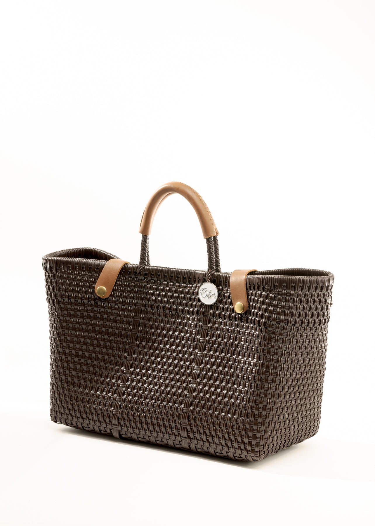 Side angle of brown woven recycled bucket handbag with brown leather handles and closure straps and a silver Coba logo medallion