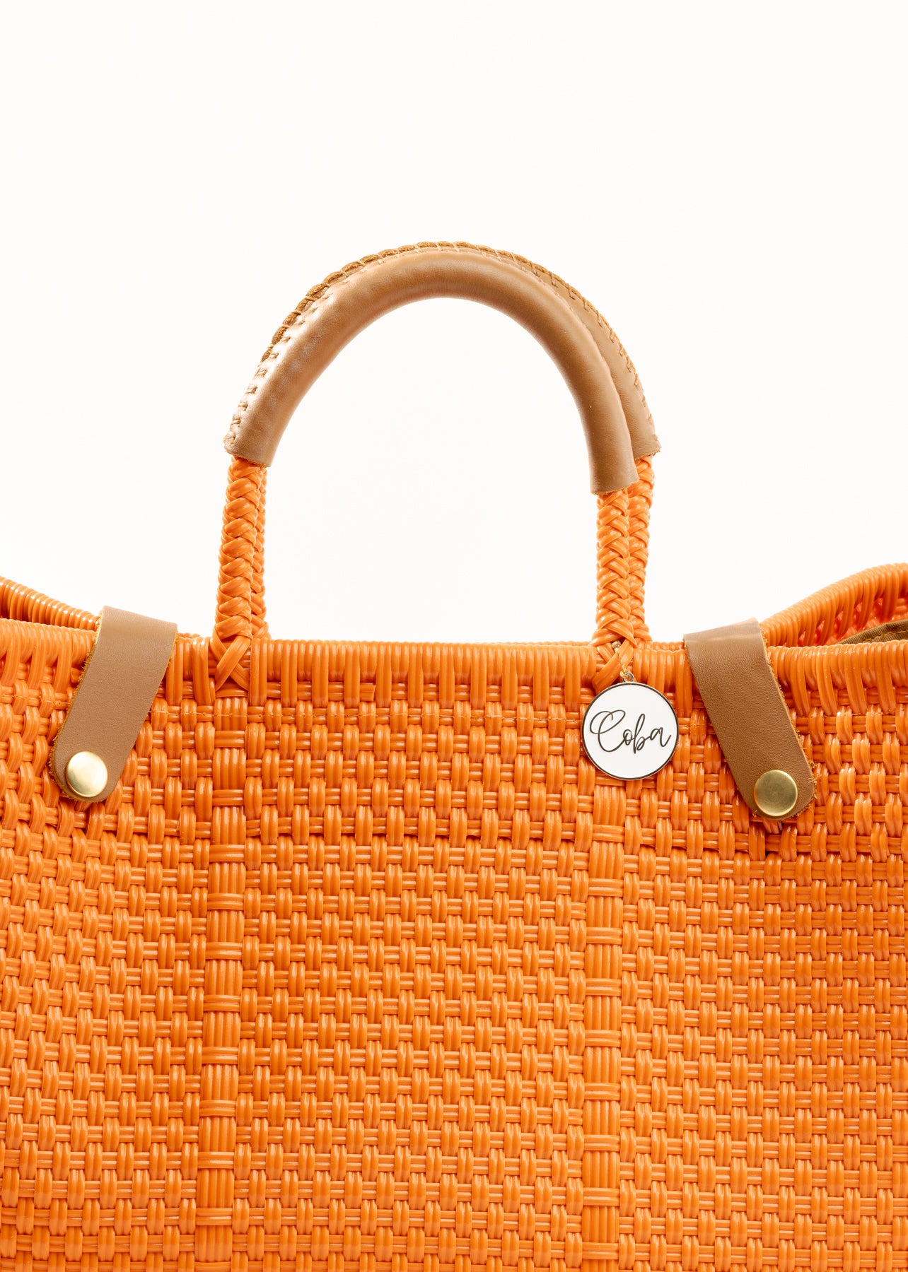 Closeup of woven fabric texture of orange woven handbag with tan leather handle, closure straps, and silver Coba medallion